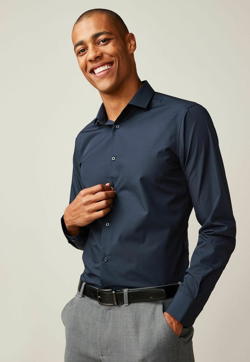 Marineblauw overhemd met knopen, kraag, gladde textuur, lange mouwen en grijze broek. Het overhemd heeft een getailleerde pasvorm en minimale details.
