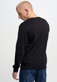 Maglione nero con maniche lunghe, scollatura a girocollo, polsini e orlo a coste, texture liscia, indossato con jeans blu, visto da dietro.