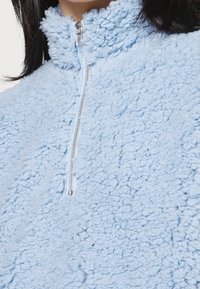 Ljusblå fleecepullover med en texturerad, plyschyta, hög krage och halvdragkedja med en silverfärgad dragkedja.
