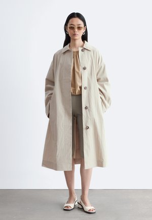 Kvinde iført beige lang frakke, tan bluse, olivengrønne knælange shorts, åbne hæle og solbriller, stående foran en ensfarvet hvid baggrund.