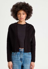 Svart cropped cardigan med ribbad textur, öppen framtill, lång ärm, kombinerad med en svart topp och ljusblå högmidjade jeans.