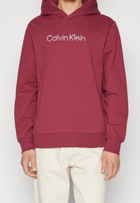 Bordowy hoodie wykonany z bawełny z przednią kieszenią typu kangur. Posiada haftowane logo "Calvin Klein" w kolorze białym. Miękka w dotyku, ze ściągaczami na mankietach.
