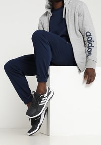 Sweat-shirt zippé gris avec le logo "adidas", joggings navy, chaussures de sport noir et blanc avec trois bandes, assis sur un cube blanc.