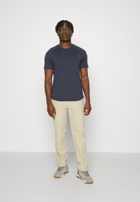 Marinefarbener Rundhalsausschnitt-T-Shirt, taillierte Passform; beige Hose. Graue Sneaker mit roten Akzenten. Einfarbiger Hintergrund, stehende Pose.