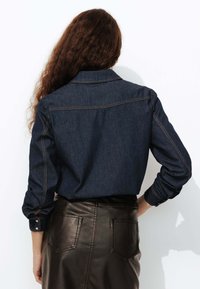 Chemise en denim foncé à manches longues et col pointu, avec des coutures contrastantes et une coupe droite. Portée avec une jupe en cuir brun.