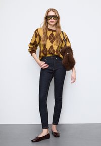 Sweter z dzianiny w żółto-brązowy wzór argyle, ciemne dopasowane dżinsy, brązowa torebka z futerkiem i brązowe płaskie buty z detalem w postaci kokardki.