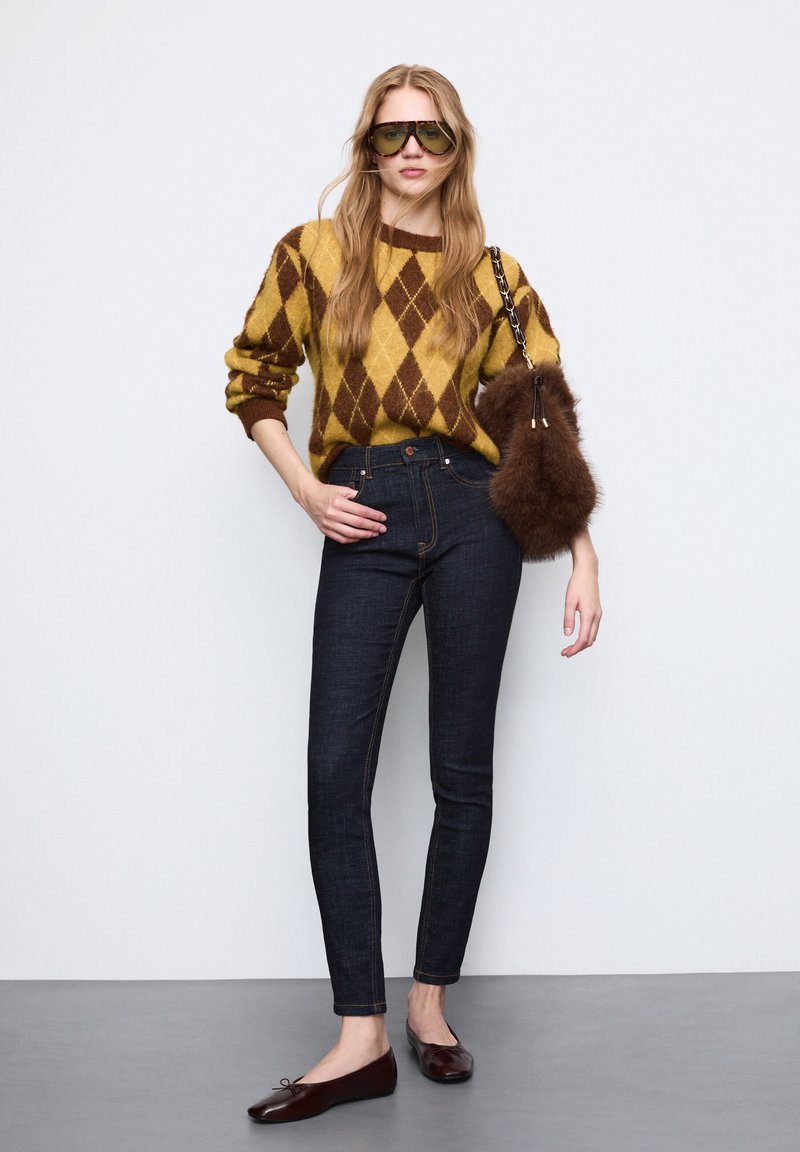 Sweter z dzianiny w żółto-brązowy wzór argyle, ciemne dopasowane dżinsy, brązowa torebka z futerkiem i brązowe płaskie buty z detalem w postaci kokardki.