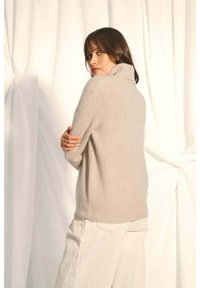 Beiger Strickpullover mit hohem Kragen, gerippten Bündchen und Saum. Er hat einen lässigen Schnitt und verfügt über abgesenkte Schulterpartien sowie Seitenschlitze.