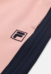 Gros plan sur un tissu rose avec une bordure côtelée bleu marine et un petit logo brodé bleu marine près de la couture d'une poche.