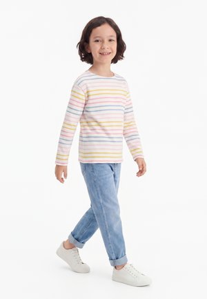 Kind mit dunklen Haaren, das ein langärmeliges pastellgestreiftes Hemd, blaue Jeans und weiße Sneakers trägt, läuft und lächelt vor weißem Hintergrund.