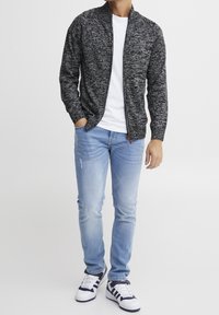 Textured black and gray zip-up cardigan, white t-shirt underneath, paired with light blue distressed jeans and white sneakers with navy accents.