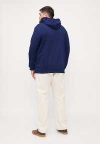 Navyblauwe hoodie met lange mouwen, een relaxte pasvorm en een kangoeroezak. Gecombineerd met crèmekleurige broeken en bruine schoenen.