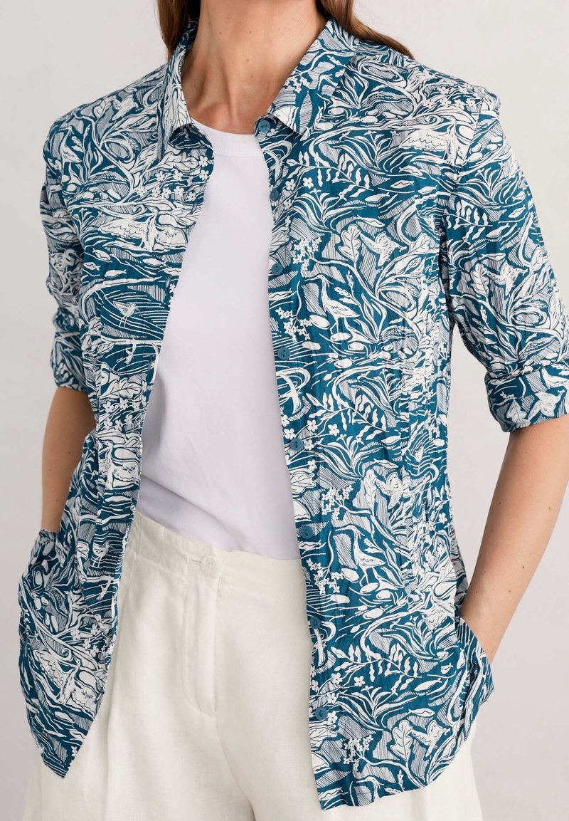 Chemise boutonnée à motif floral teal et blanc, dotée de manches trois-quarts, d'un col et de deux poches avant.