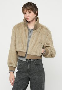 Chaqueta bomber de piel sintética beige con puños y cuello acanalados. Cuenta con cierre de cremallera y bolsillo lateral. Llevado sobre una blusa gris y jeans negros.