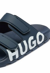 Marina slide sandals med kardborreband, texturerad gummisula och stor vit "HUGO"-bokstäver på sidan, designade för komfort och avslappnad användning.