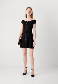 Robe noire décolletée, avec des boutons sur le devant, un design plissé et un col festonné. Portée avec des chaussures à talons noirs et une pochette.