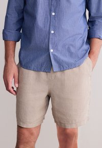 Pantaloncini in lino beige chiaro con vestibilità rilassata. Abbinati a una camicia a righe blu con collo e bottoni bianchi.