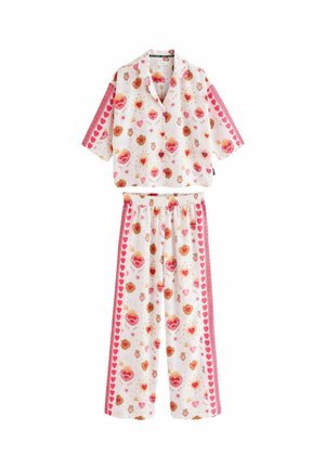 Vrouw draagt losse crèmekleurige pyjama met rood hart- en zonpatroon, staat met een hand achter het hoofd tegen een neutrale muur.