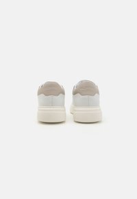 White leather sneakers with beige accents, featuring a padded collar, textured outsole, and logo detail on the heel.