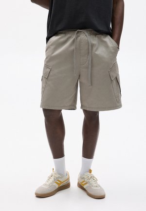 Beige Cargo-Shorts mit elastischem Bund und Kordelzug, ausgestattet mit zwei Seitentaschen und einem lockeren Schnitt. Getragen mit weißen Socken und Sneakers.