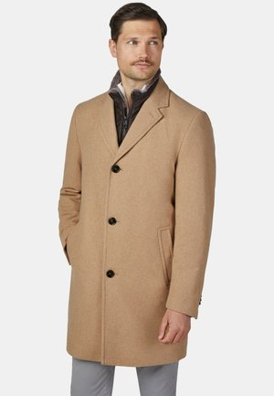 Mann trägt einen hellbraunen Wollmantel mit drei Knöpfen über einer dunklen Reißverschlussjacke, steht mit einer Hand in der Tasche und schaut zur Seite.