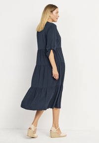 Mörkblå, lager-på-lager klänning med trekvartsärmar, knapppdetaljer och en flödande nederkant, ihop med beige espadrillosandaler.