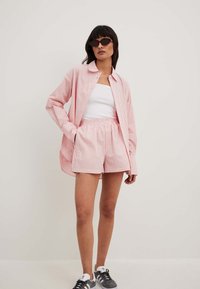 Rožinė ir balta dryžuota marškiniai, derinami su atitinkančiomis šortais, pagamintais iš lengvos medžiagos. Modelis dėvi juodus akinius nuo saulės ir pilkus sportbačius.