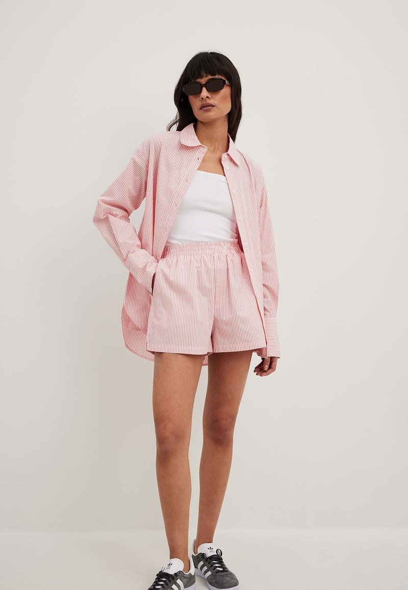 Rožinė ir balta dryžuota marškiniai, derinami su atitinkančiomis šortais, pagamintais iš lengvos medžiagos. Modelis dėvi juodus akinius nuo saulės ir pilkus sportbačius.