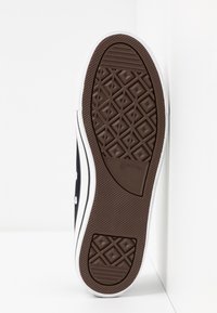 Semelle d'une basket Converse All Star noire et blanche montrant une semelle en caoutchouc marron avec des motifs en losange et un logo au centre.