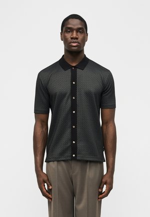 Mees, kes kannab musta lühikeste varrukatega mustrilist nööbitavat särki musta kraega ja beeže pükse, seisab ühevärvilise heleda tausta ees.