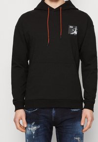 Černá mikina s kapucí, přední kapsou, nastavitelnou kapucí se červenými stahovacími šňůrkami a kontrastní nášivkou loga na hrudi. Smyčková látka z bavlny.
