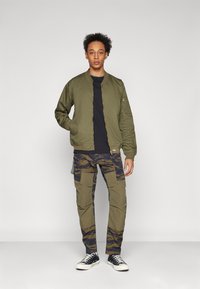 Chaqueta bomber verde oliva con cremallera frontal, combinada con pantalones cargo que presentan un estampado de camuflaje azul y múltiples bolsillos. Las zapatillas negras completan el look.