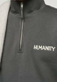 Sweat-shirt gris avec un col haut, une demi-fermeture éclair et "HUMANITY" brodé en blanc. Texture lisse avec un design simple et moderne.