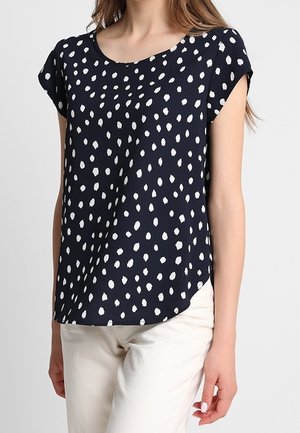Donna che indossa una blusa a maniche corte blu navy con macchie bianche irregolari e pantaloni color crema, in piedi davanti a uno sfondo neutro.
