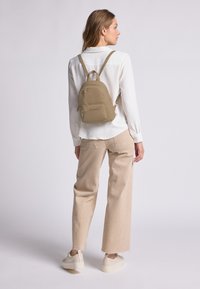 Mochila marrón con un acabado texturizado, que cuenta con un bolsillo frontal con cremallera y correas ajustables, llevada sobre una camisa blanca y pantalones beiges.