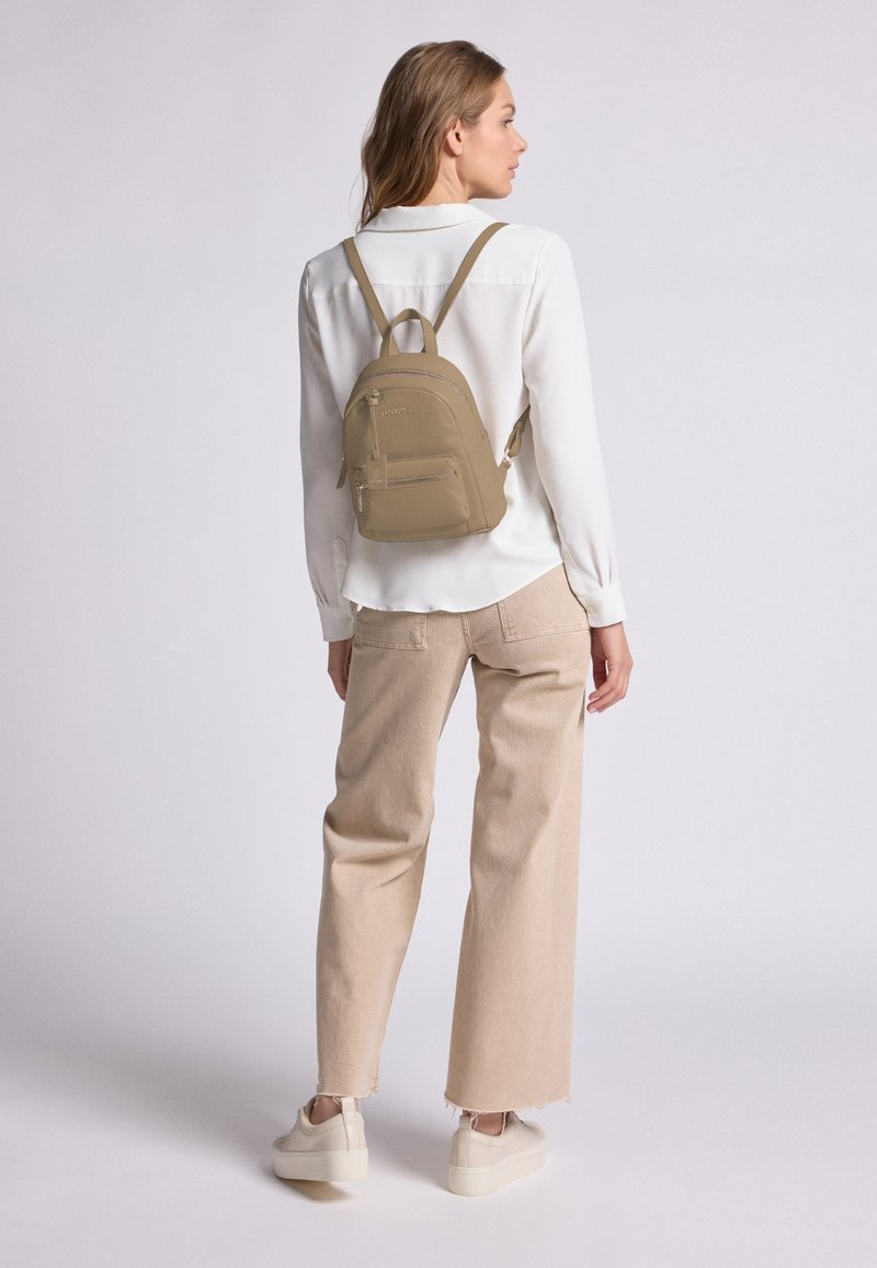 Mochila marrón con un acabado texturizado, que cuenta con un bolsillo frontal con cremallera y correas ajustables, llevada sobre una camisa blanca y pantalones beiges.