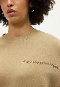 Béžová mikina s kulatým výstřihem, vyrobená z hladkého materiálu, s jemným dekorativním textem v tmavě hnědé barvě umístěným na přední straně.