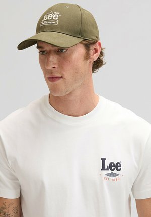 Casquette de baseball kaki avec logo "Lee" brodé en blanc et visière incurvée, portée avec un t-shirt blanc uni arborant un petit logo sur la poitrine.