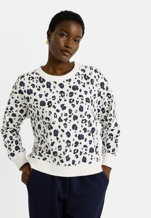Femme portant un sweatshirt blanc à manches longues avec un imprimé léopard noir et bleu, assorti à un pantalon bleu foncé, se tenant avec les mains dans les poches.