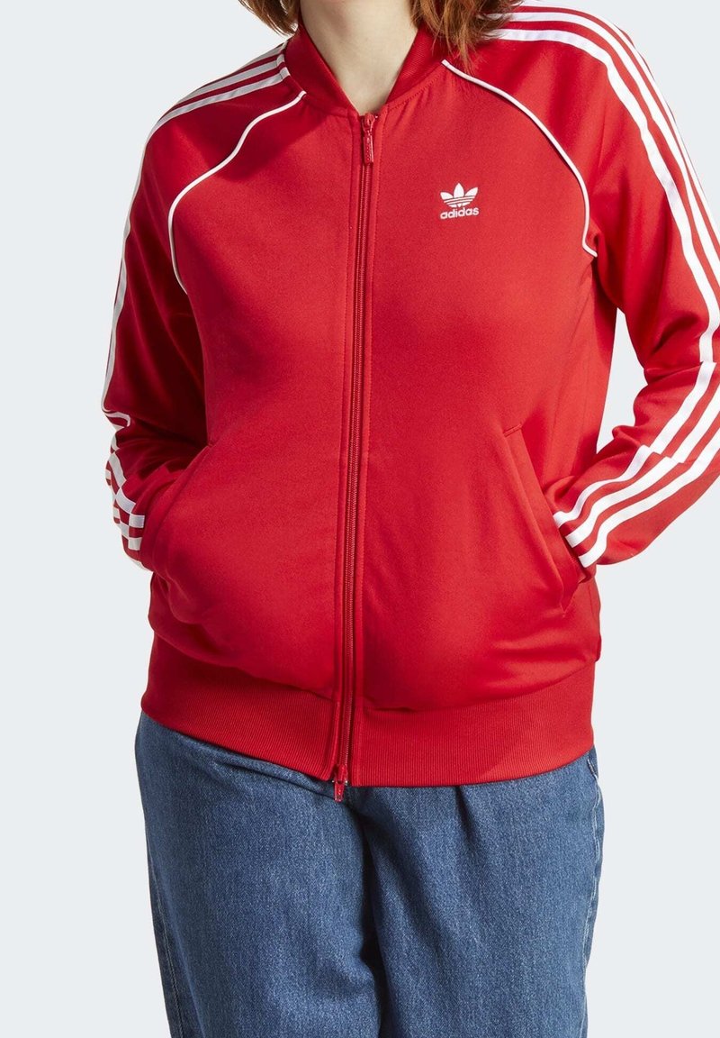 Červená bunda na zip z hladké látky, s bílými pruhy na rukávech a malým logem na hrudi. Přední kapsy dvě.