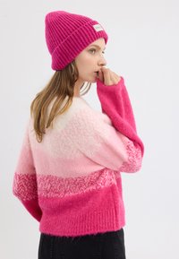 Rosa und fuchsia Pullover mit einem Farbverlauf-Design, ausgestattet mit lockerer Passform und weiten Ärmeln. Eine passende gerippte rosa Mütze rundet das Ensemble ab.