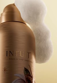 Flacon en spray en bronze avec une finition texturée. Présente de la mousse blanche émergeant du dessus. L'étiquette affiche "INTUITIA" et un design floral.