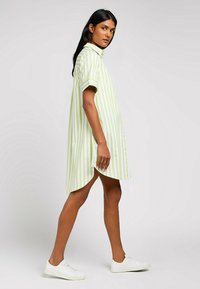 Abito a camicia con bottoni di colore verde chiaro a righe bianche, colletto, maniche corte, fessure laterali, realizzato in tessuto leggero, abbinato a sneaker bianche.
