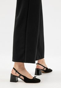 Décolleté neri con un texture vellutato, tacco grosso e lucido, e un'accattivante decorazione a fiocco sulla punta. Abbinati a pantaloni larghi neri.