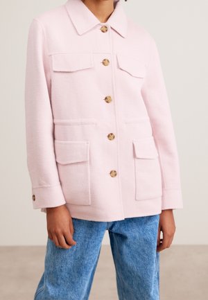 Personne portant une veste rose clair boutonnée avec quatre poches à l'avant et un jean en denim bleu, debout à l'intérieur.
