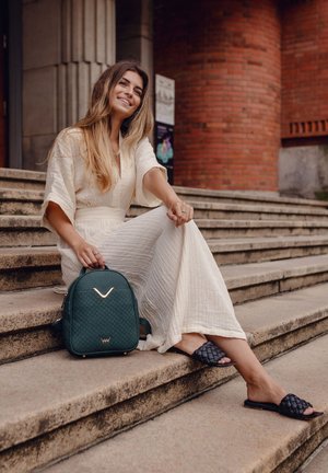 Green quilted backpack with gold hardware, worn on steps. The model wears a cream dress and black woven slide sandals.