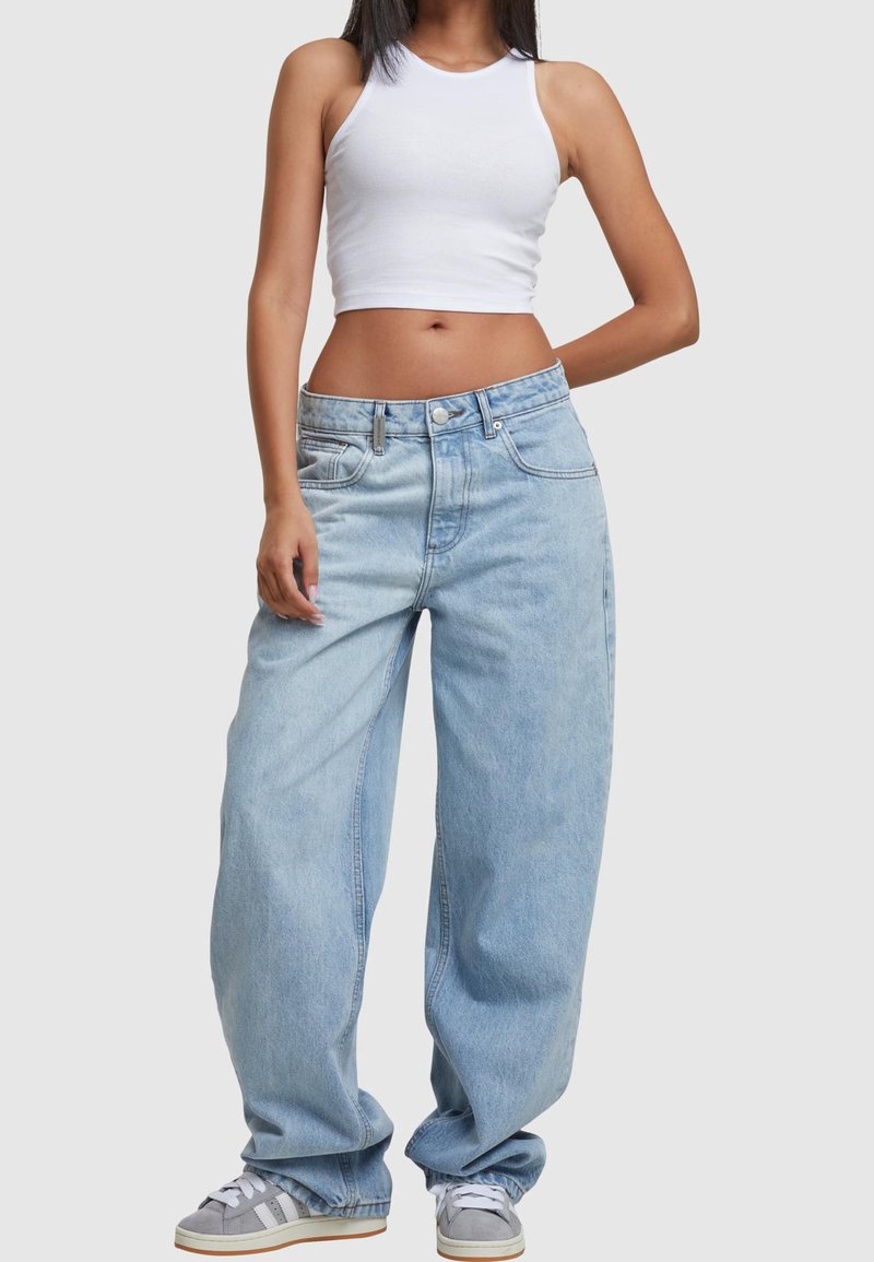 Femme portant un crop top blanc sans manches, un jean bleu clair ample et des baskets grises, debout devant un fond uni.