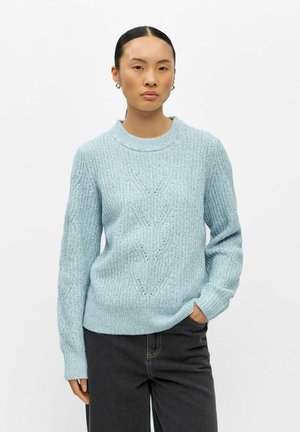 Femme aux cheveux foncés portant un pull en maille bleu clair et un jean noir, posant avec une main dans la poche devant un fond blanc uni.