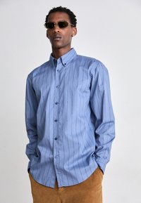 Camicia a maniche lunghe azzurro chiaro con righe bianche verticali e una texture liscia, abbinata a occhiali da sole con lenti color oliva e pantaloni in velluto a coste marroni.