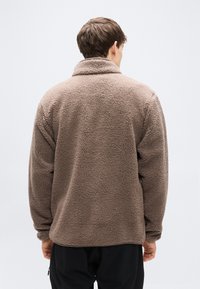 Καφέ φλις μπουφάν με ψηλό γιακά, άνετη εφαρμογή και υφή επιφάνειας. Διαθέτει μακριά μανίκια και ίσιο τελείωμα.